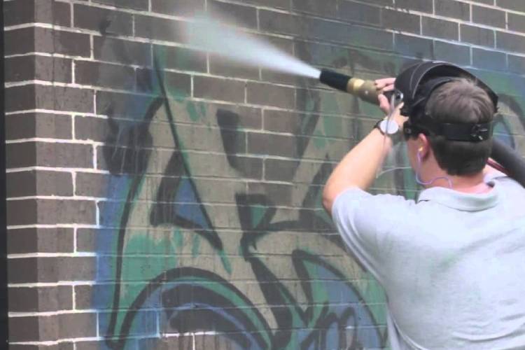 A professional technician using a high-powered dustless blasting machine to remove graffiti from a brick wall. The blasting nozzle sprays a fine mist at the graffiti-covered surface, visibly stripping away paint while leaving the clean brick underneath. The technician is wearing protective gear, including a face shield and ear protection, ensuring safety during the graffiti removal process.