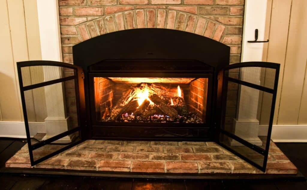 A cozy gas fireplace insert burning with warm flames, set in a traditional brick fireplace with black metal doors open, showcasing the inviting ambiance of a converted wood-burning fireplace.