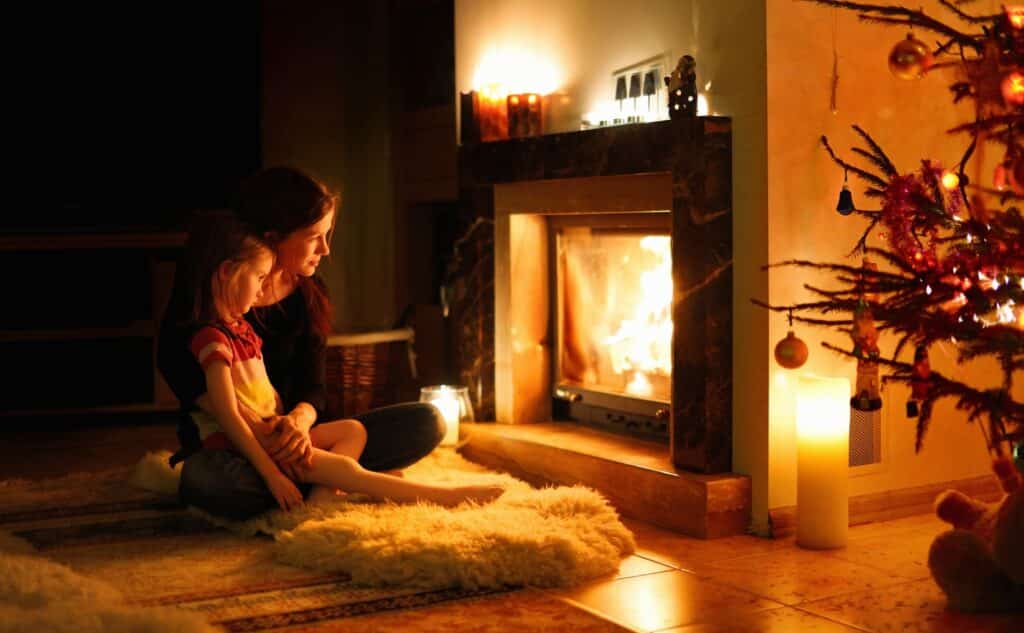 A mother and her young daughter sit together on a cozy rug near a warm fireplace, watching the fire safely behind a glass door. The room has a festive atmosphere with a softly lit Christmas tree and candles, creating a peaceful and family-friendly setting. This image represents the importance of fireplace safety for children, ensuring a secure and comfortable home environment.