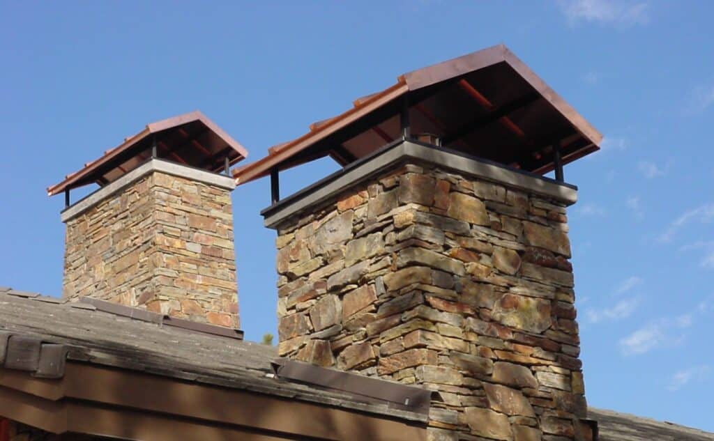 Stone chimneys with protective metal chimney caps under a clear blue sky. Installing a chimney cap helps prevent debris, animals, and moisture from entering your chimney, enhancing safety and efficiency.