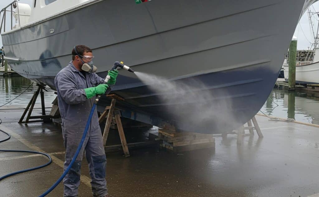 A professional technician performing Dustless Blasting on a fishing boat hull at a marina, safely removing old marine paint using an eco-friendly, dust-free method.