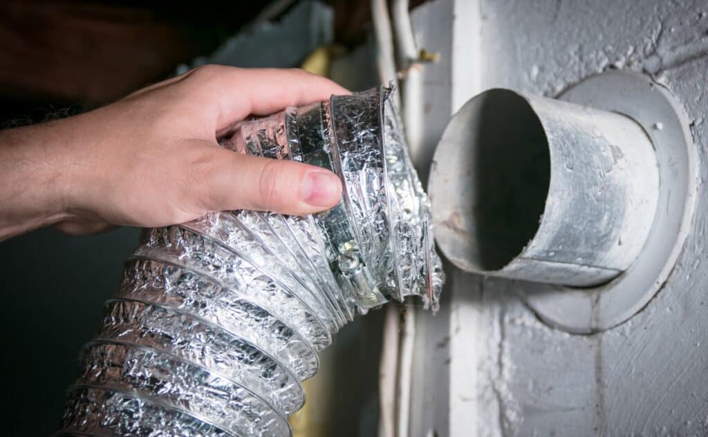 A close-up of a hand connecting a flexible aluminum dryer vent hose to a wall duct, emphasizing proper dryer vent maintenance. The metal duct shows signs of dust and lint buildup, highlighting the need for regular cleaning to ensure safety and efficiency. This image reinforces the importance of professional dryer vent cleaning in preventing fire hazards and improving appliance performance.